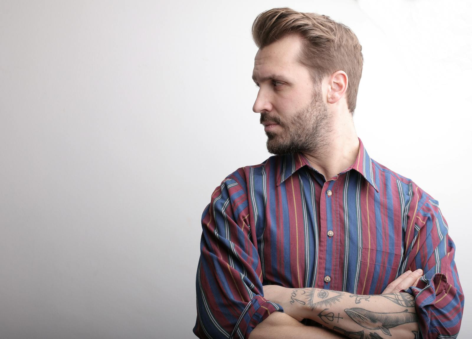 Portrait of a bearded man with tattoos looking to the side in a striped shirt.