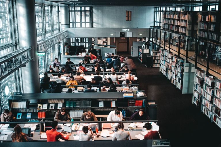 From above of anonymous students with netbooks and books preparing for exams near bookshelves with literature collection in university library